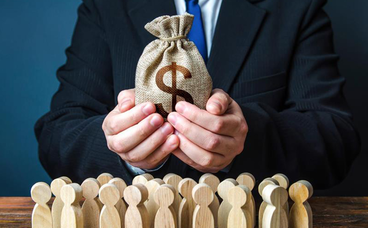 A person in a suit holds a burlap sack with a dollar sign in front of wooden figurines.