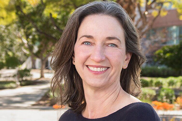 Smiling woman, American Studies Professor Carrie Lane, in an outdoor setting with trees and flowers in the background.