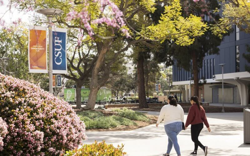 CSUF campus with people walking on a path, flowering bushes, and banners displaying "CSUF."