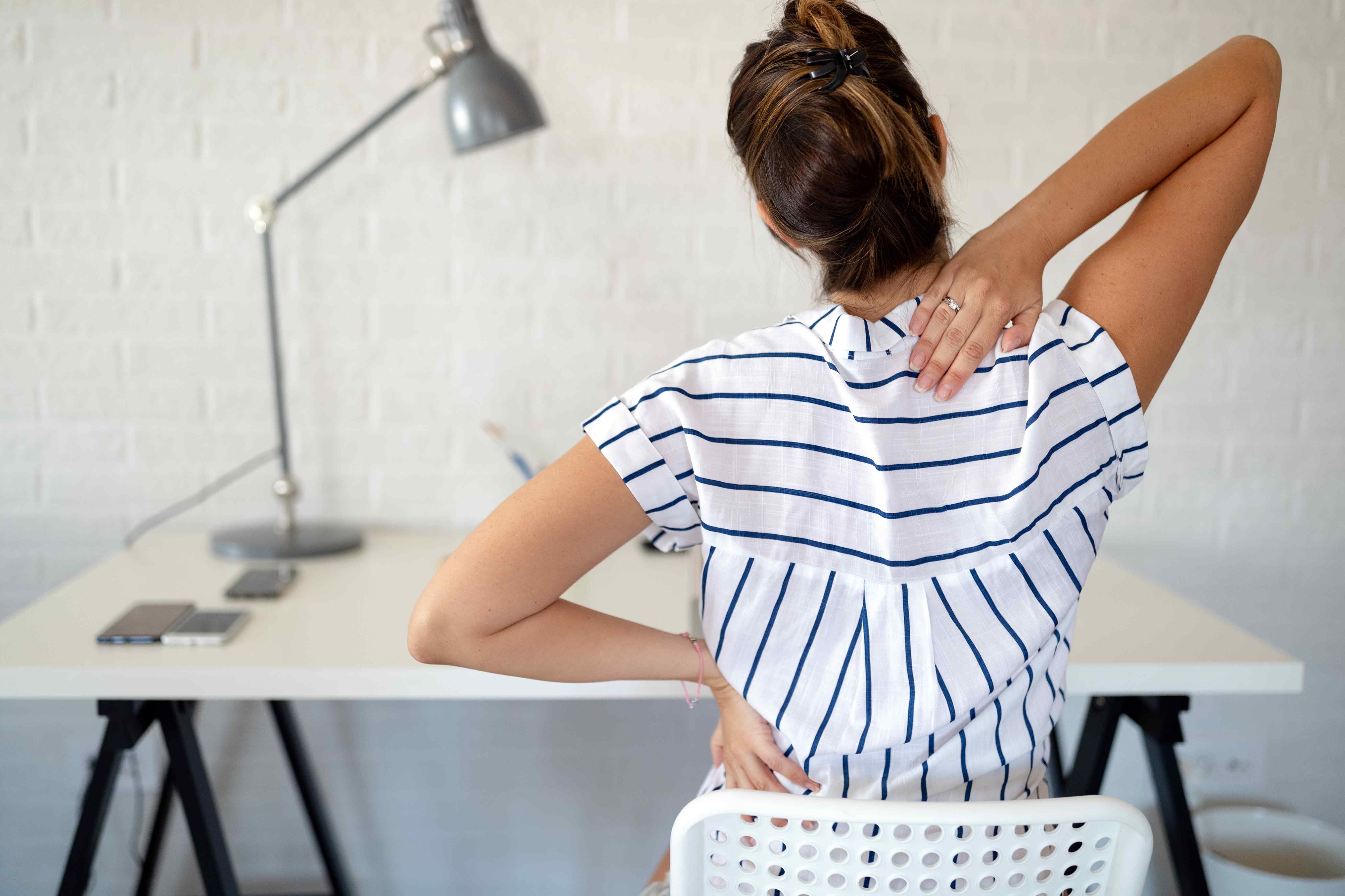 Person sitting at a desk holding their neck and lower back, suggesting discomfort.