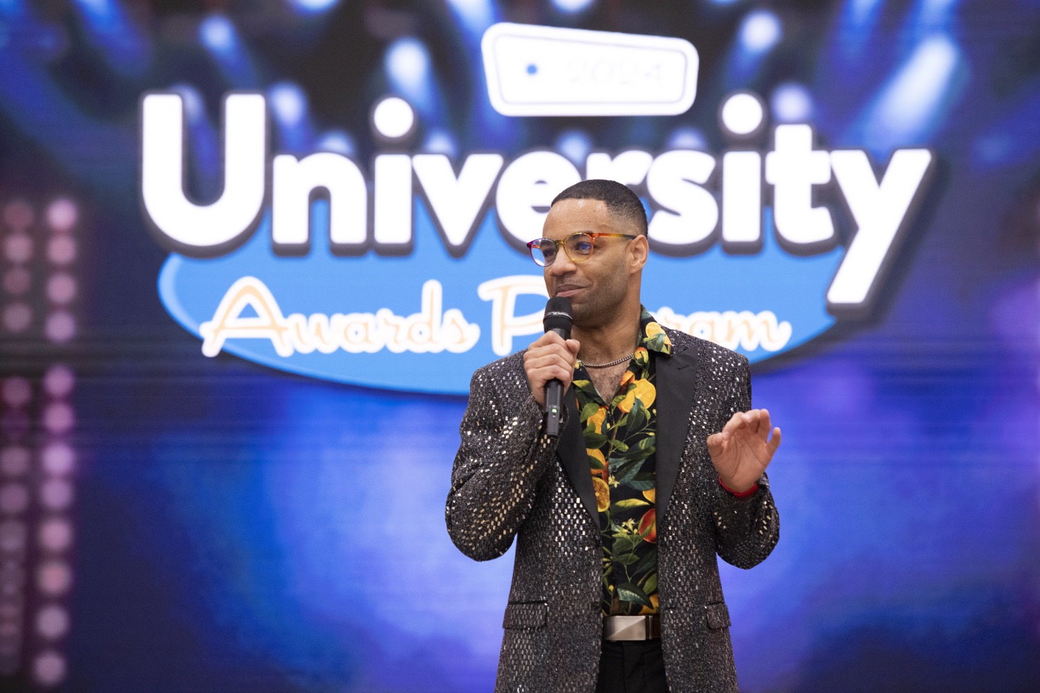 UAP MC Tim Alexander holding a microphone, wearing a sparkly blazer and floral shirt, standing in front of a "University Awards Program" backdrop